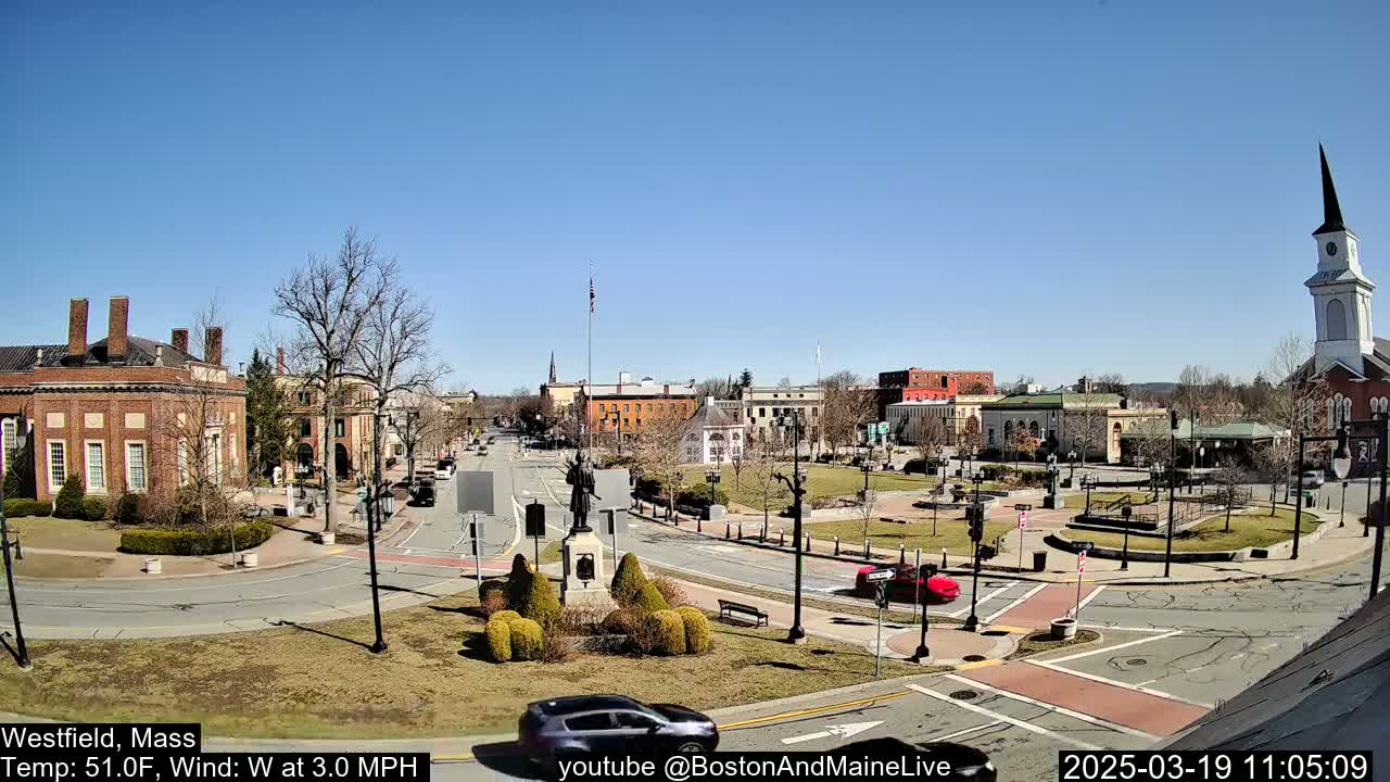 A sunny town square with brick buildings, a monument, and sparse traffic under a clear blue sky.