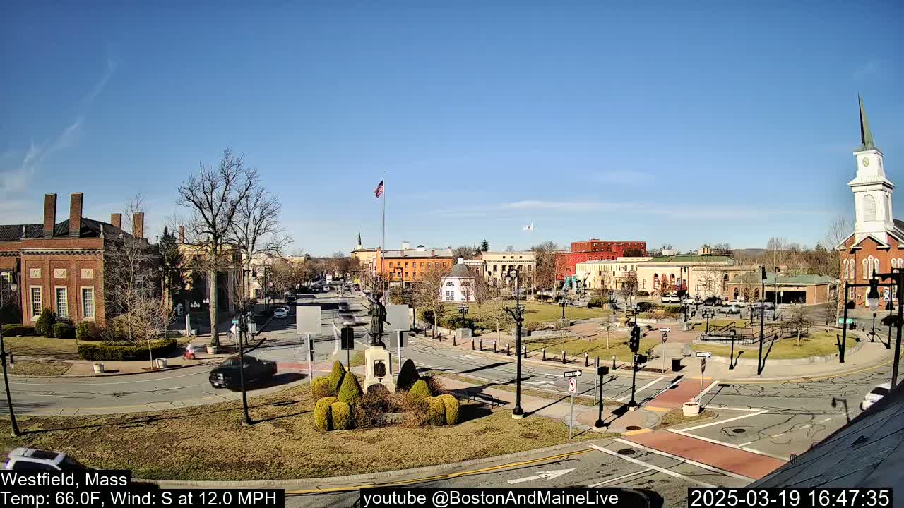 A town square with brick buildings, a statue, and a church steeple under a clear blue sky and sunny conditions.