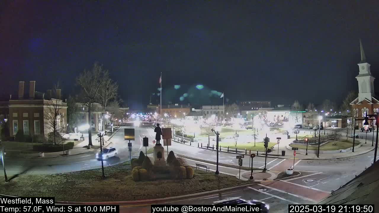 A nighttime view of a town square with a statue in the center, surrounded by buildings, roads, and sparse traffic under a clear, dark sky with a south wind.