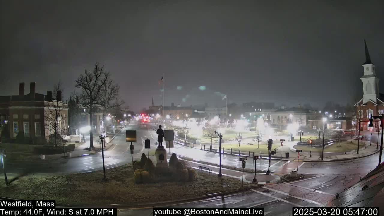 A nighttime view of a town square with a statue, several buildings, and wet roads under a dark, overcast sky.