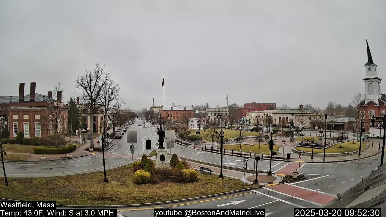 A mostly cloudy town square with a statue in the center, surrounded by brick buildings and a church steeple in the background.