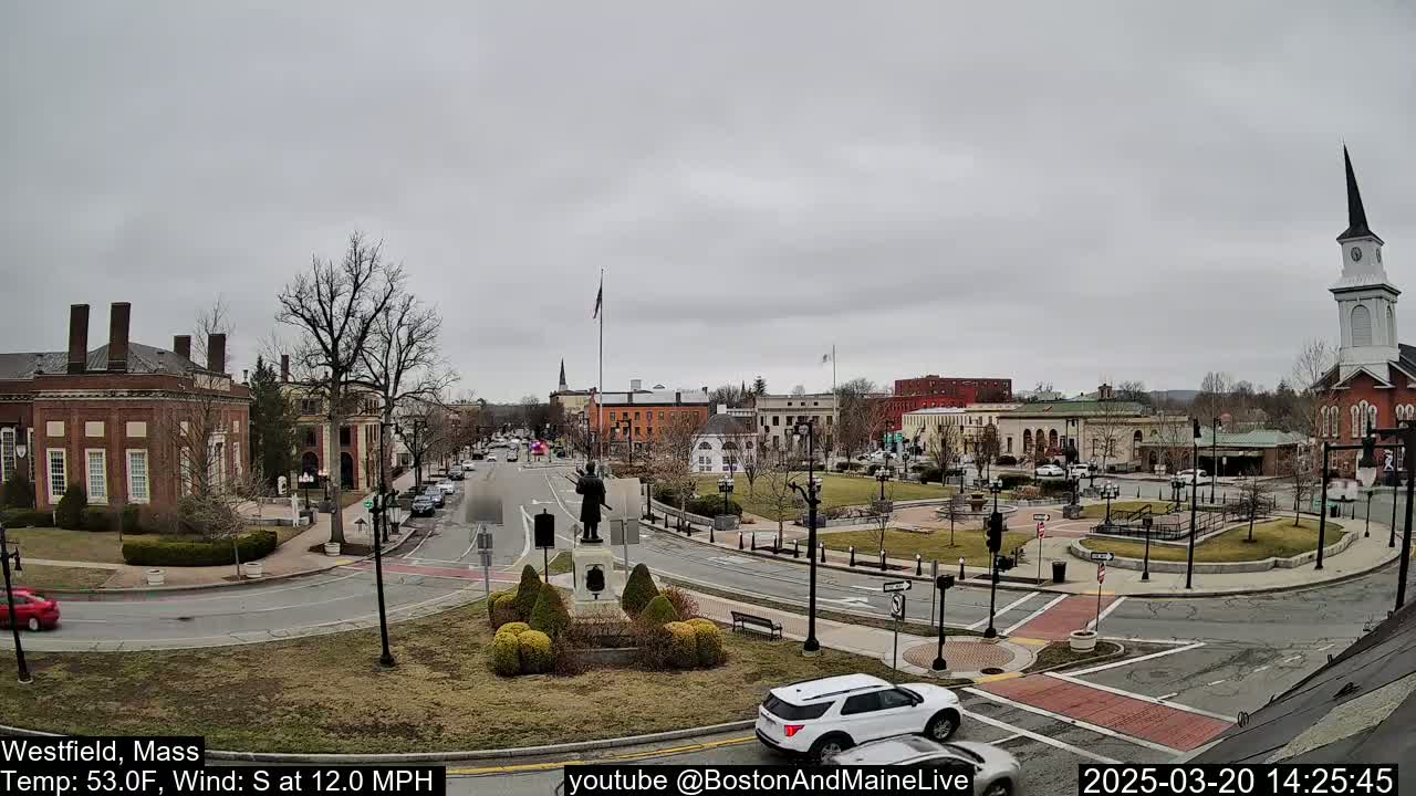 A town square with several brick buildings, a statue, and sparse traffic under an overcast sky.