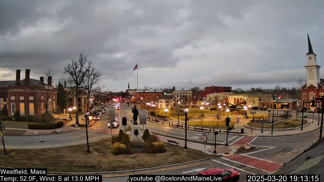 A town square with brick buildings, a statue, and a church steeple under a cloudy sky, with a moderate south wind.