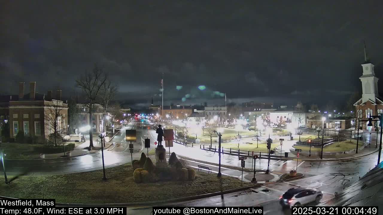 A nighttime view of a town square with wet roads, a statue in the center, and a few cars driving by under a cloudy sky.