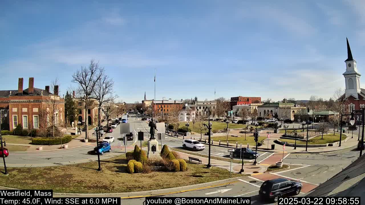 A sunny day in a town square shows brick buildings, a statue, cars, and a church steeple under a clear blue sky with a light wind.