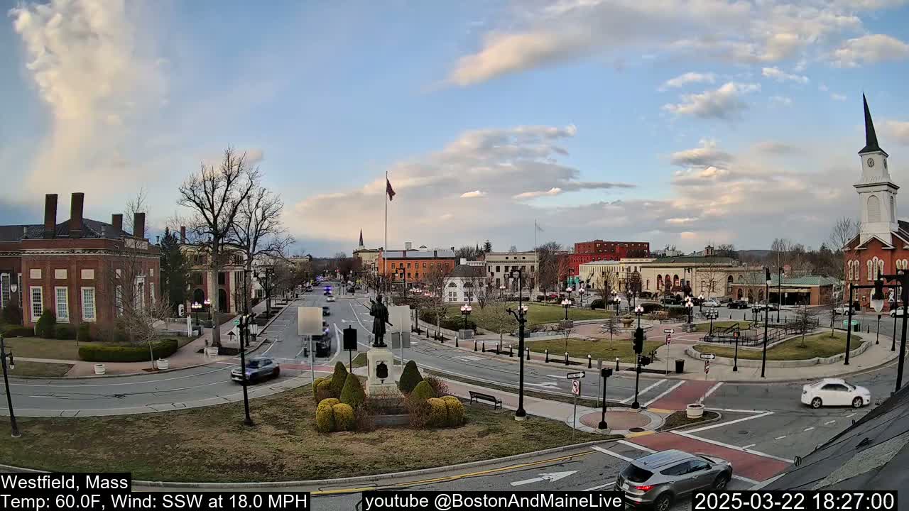 A partly cloudy town square with brick buildings, a monument, and cars driving on roads under a mostly clear sky.