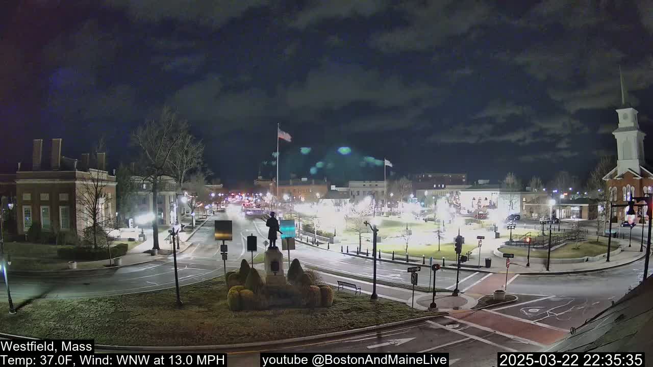 A nighttime view of a town square with a statue in the center, under a partly cloudy sky, with a 13 mph wind.
