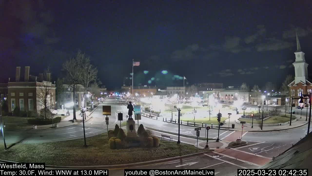 A nighttime view of a town square with a statue in the center, under a partly cloudy sky with a wind blowing from the west-northwest at 13 mph.