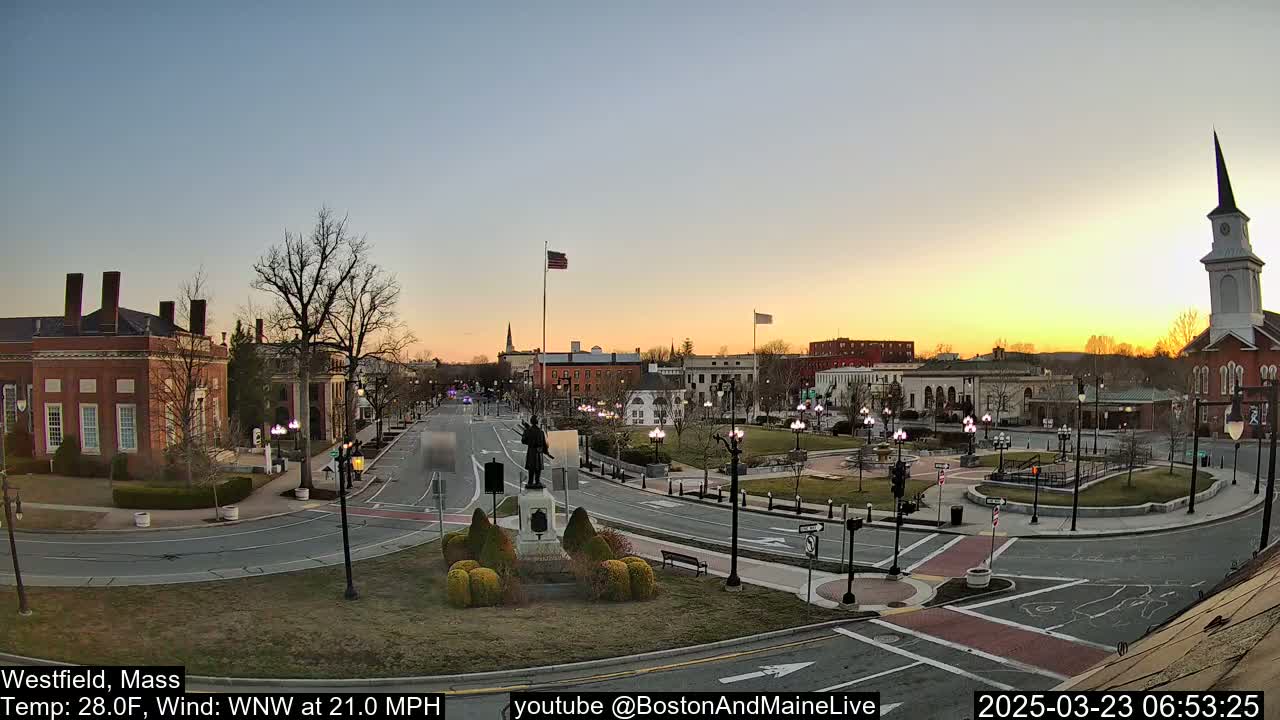 A town square at sunset, with a statue in the center, surrounded by brick buildings and a church steeple under a clear sky and a brisk wind.