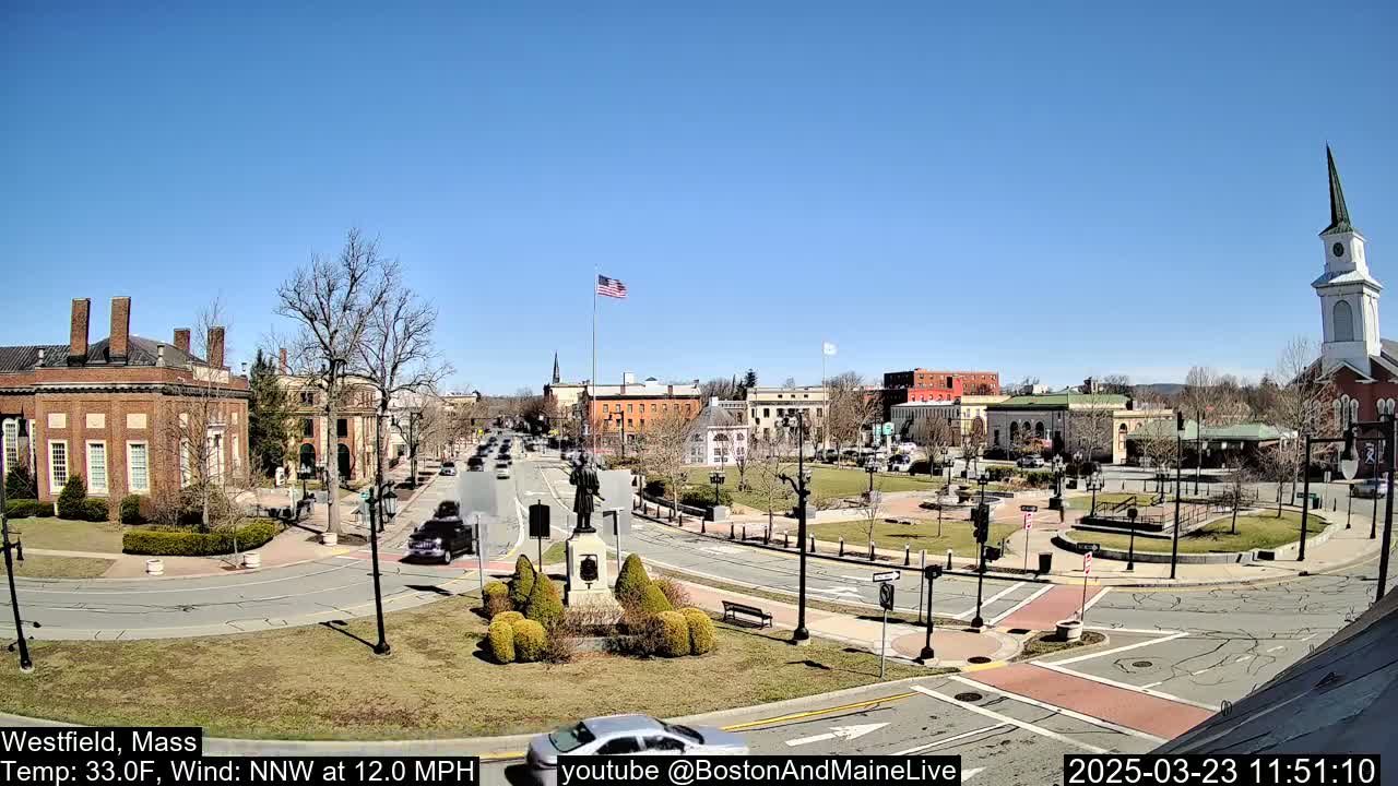 A town square with brick buildings, a monument, and a church steeple under a clear blue sky.