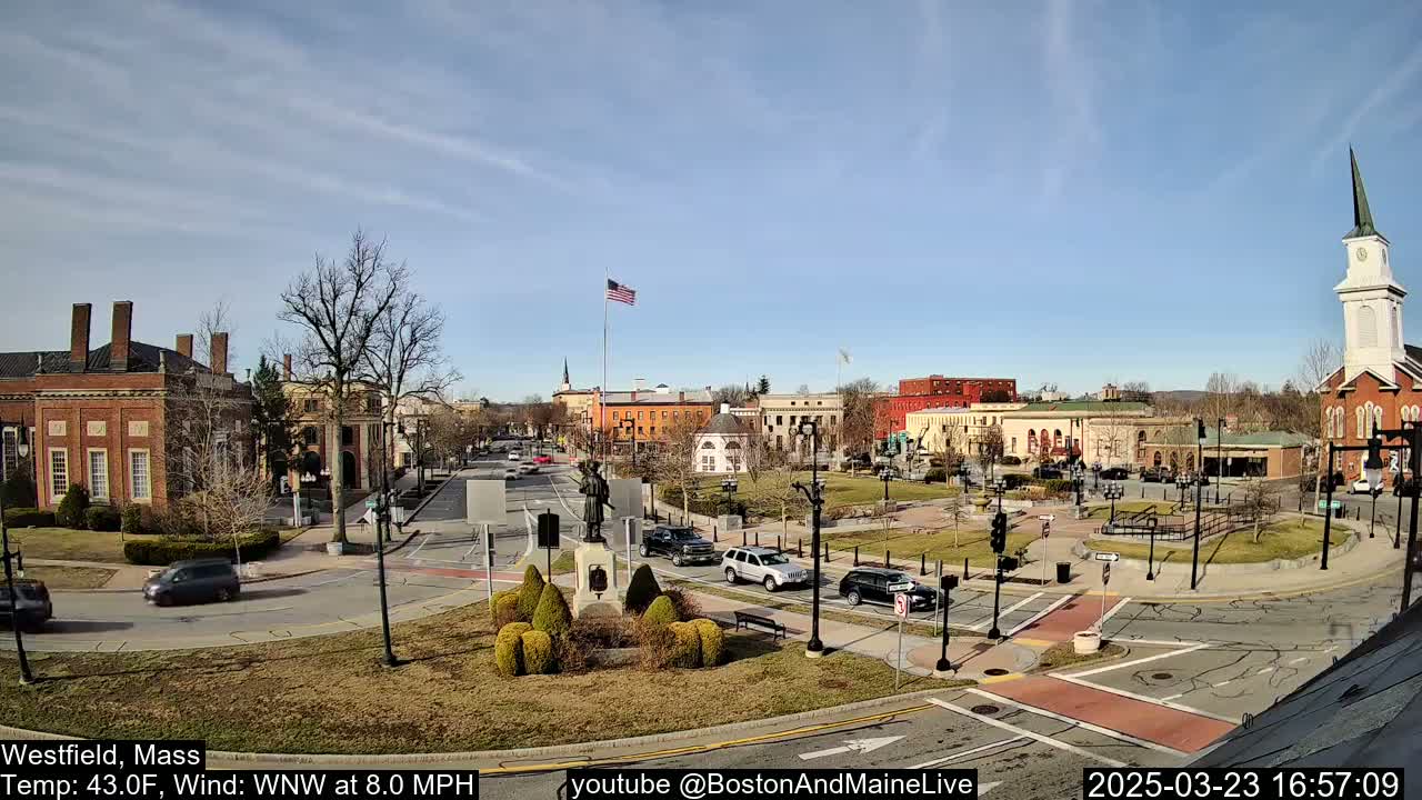 A town square with brick buildings, a monument, and a church steeple under a partly cloudy, sunny sky.