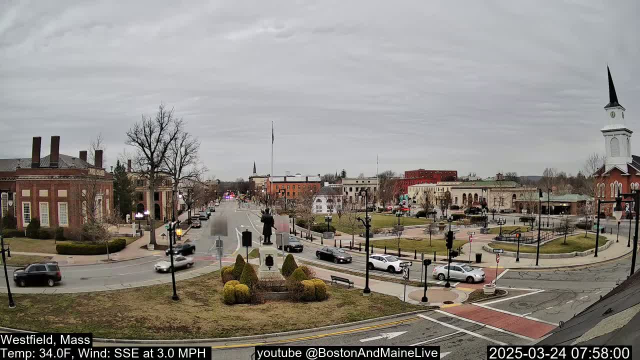 A town square with brick buildings, a statue, and sparse traffic under an overcast sky.