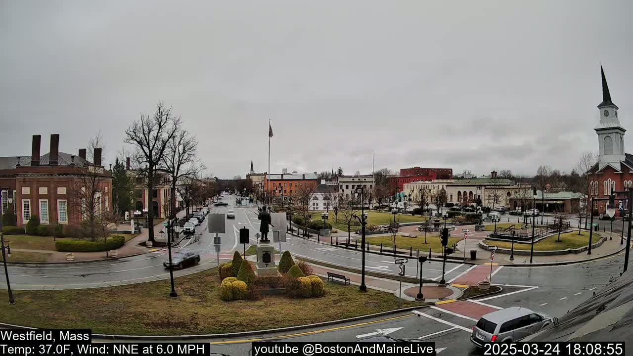 A town square with brick buildings, a statue, and sparse traffic under an overcast sky.