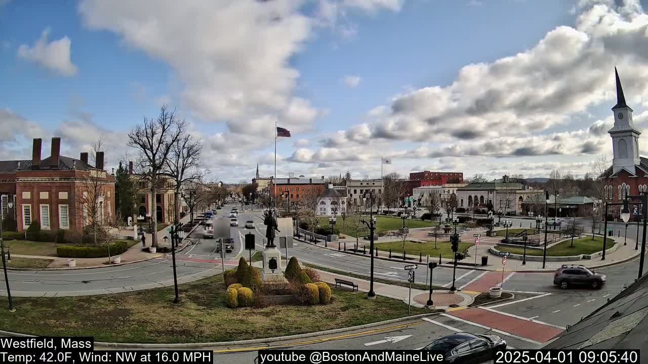 A partly cloudy day shows a town square with a monument in the center, brick buildings, a church steeple, and light traffic on the roads.