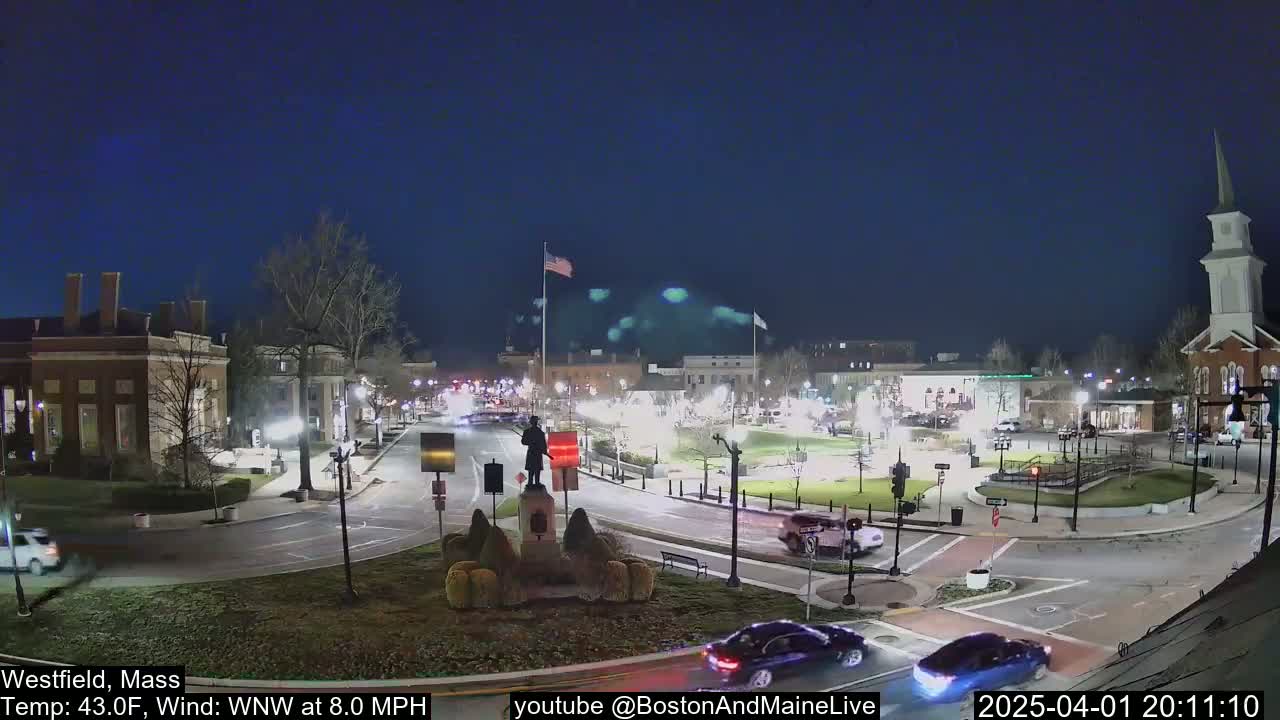 A nighttime view of a town square with a statue in the center, surrounded by buildings and cars, under a clear, dark sky with a light wind.