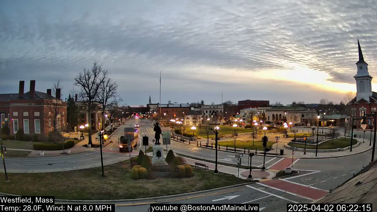 A town square with a statue in the center, surrounded by buildings and a church steeple, at sunset under a partly cloudy sky.