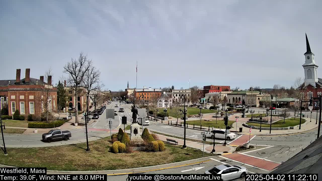 A town square with brick buildings, a statue, and sparse traffic under a mostly clear sky.