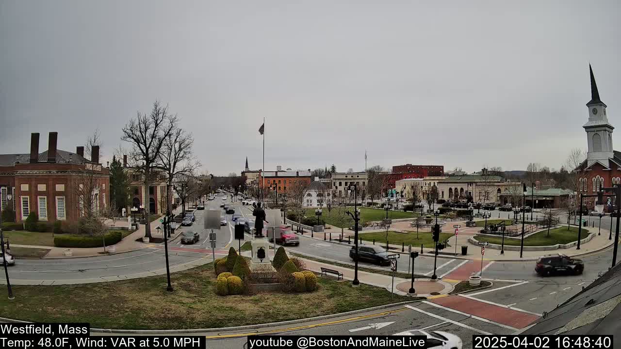 A town square with brick buildings, a statue, and sparse traffic under an overcast sky.