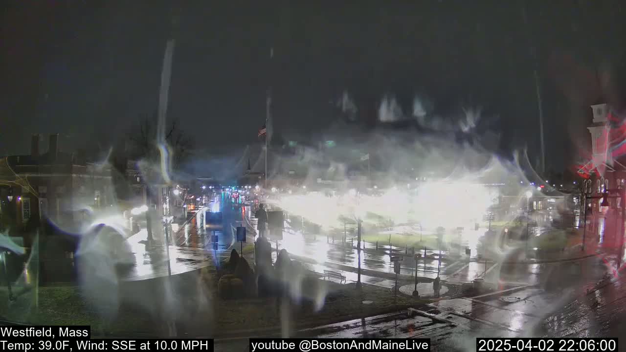 A rain-swept nighttime scene shows a town square with wet streets and vehicles, under a dark sky with streaks of what appears to be heavy spray or possibly snow.