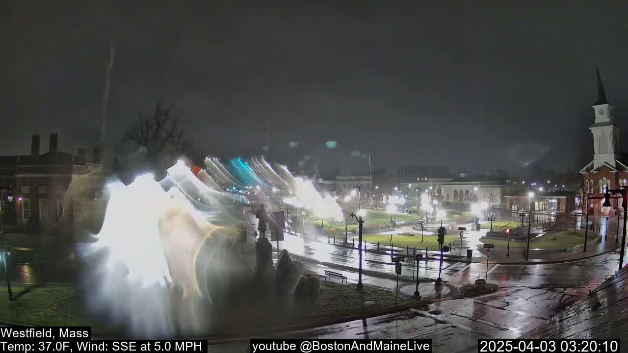 A nighttime view of a town square with a church steeple in the background, wet streets, and light streaks possibly from rain or camera lens flare, under a dark, overcast sky.