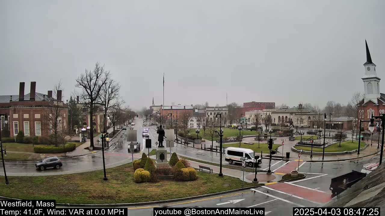 A wet town square with a statue in the center, surrounded by brick buildings and light traffic under a gray, overcast sky.