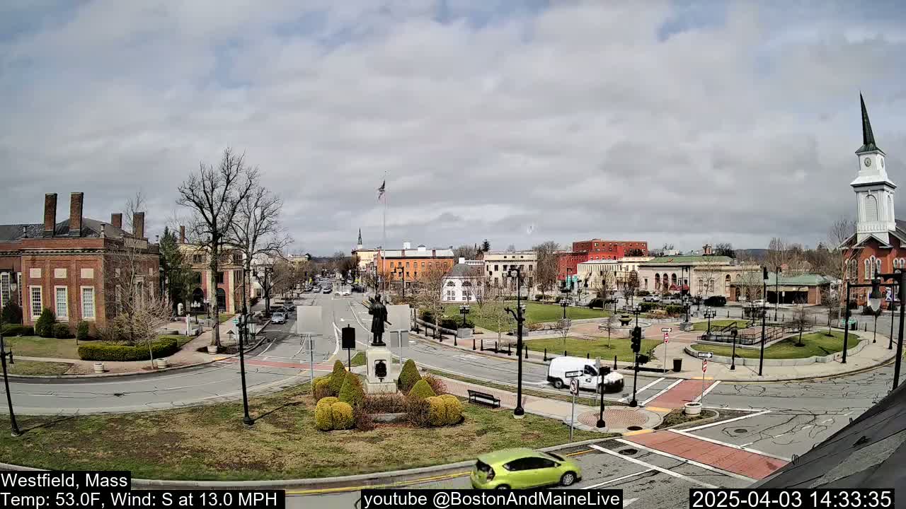 A town square with brick buildings, a monument, and sparse traffic under a partly cloudy sky.