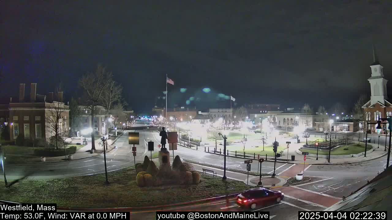 A nighttime view of a town square with a statue in the center, surrounded by buildings and roads, under a clear, dark sky.