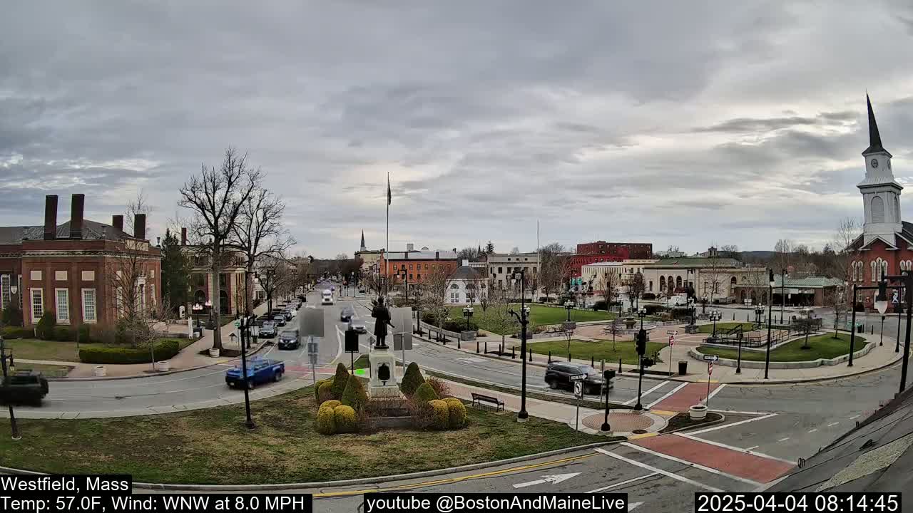 A partly cloudy day reveals a town square with a statue at its center, surrounded by brick buildings and cars on roads.