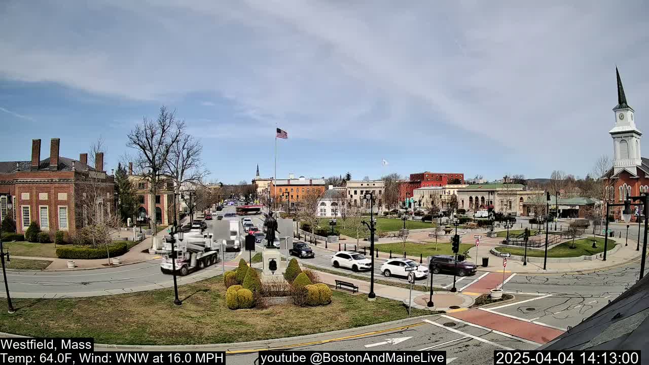 A partly sunny day shows a town square with several brick buildings, a monument, and cars driving around a roundabout.