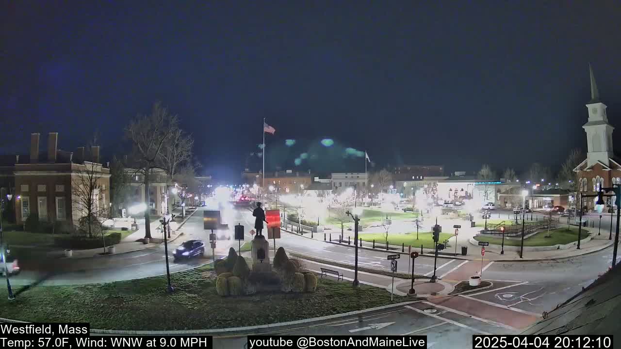 A nighttime view of a town square with a statue, several buildings, and sparse traffic under a clear, dark sky.