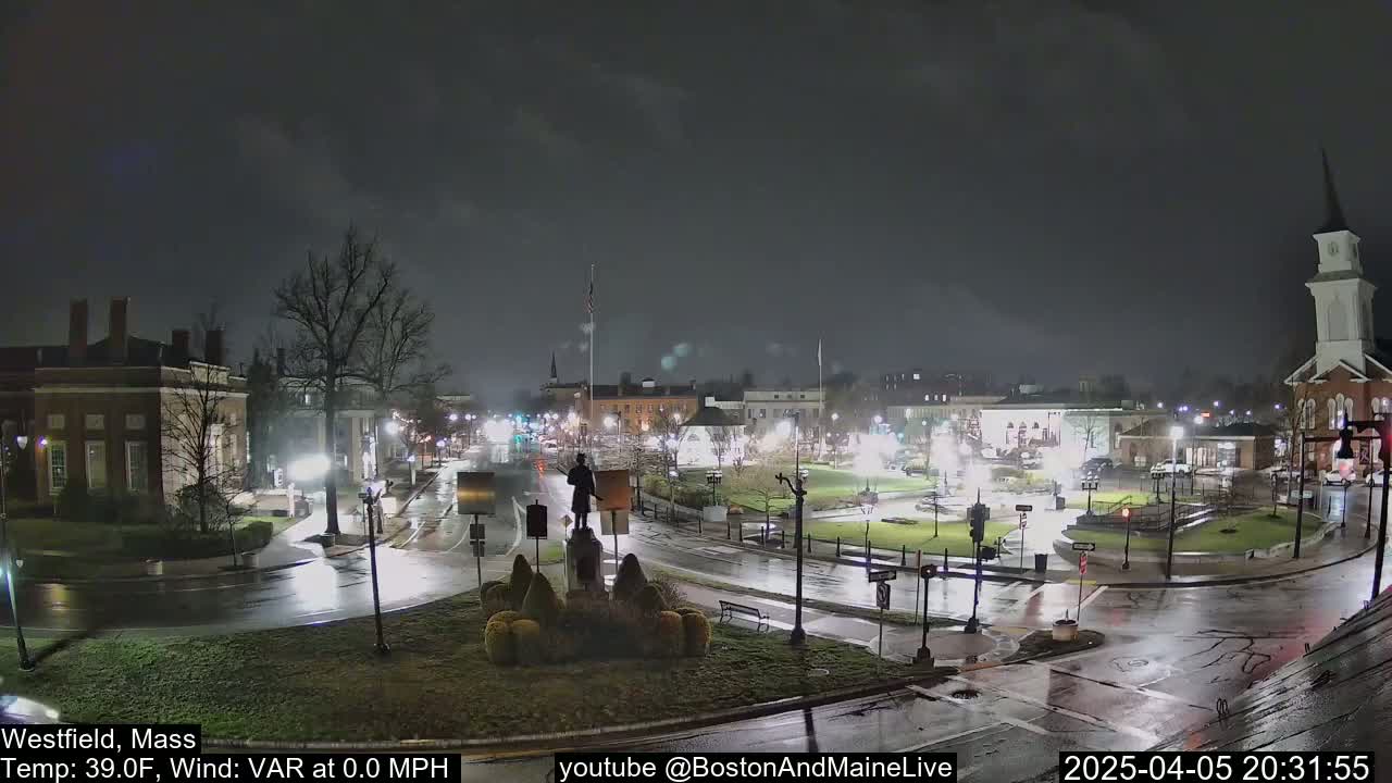 A nighttime, rain-slicked town square with a statue at its center, surrounded by buildings and streetlights.