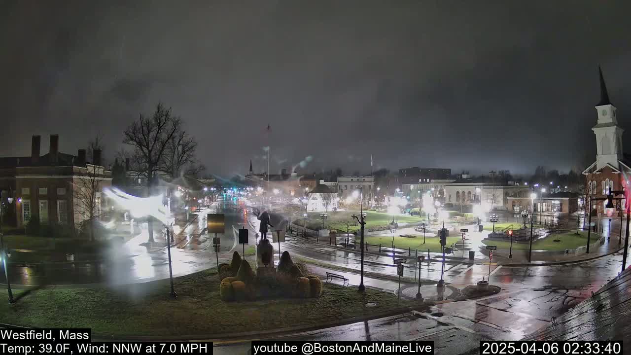 A nighttime view of a town square with wet streets and a church steeple under a dark, overcast sky.