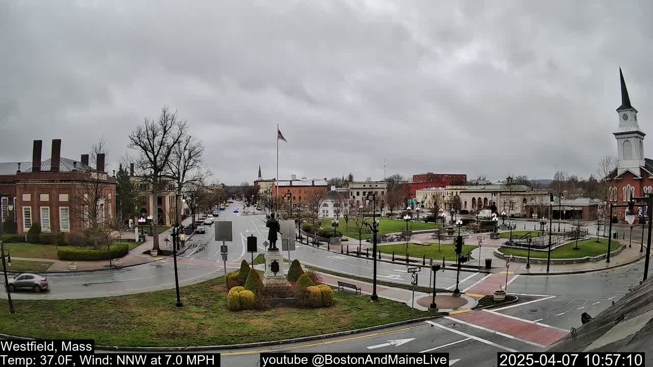 A town square with several brick buildings, a statue, and a church steeple under an overcast sky.