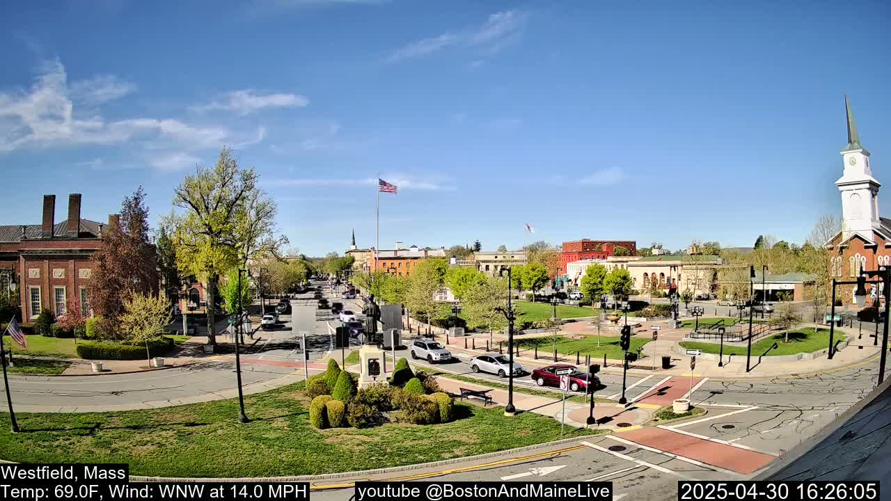 A town square with brick buildings, green spaces, and cars under a clear, sunny sky.