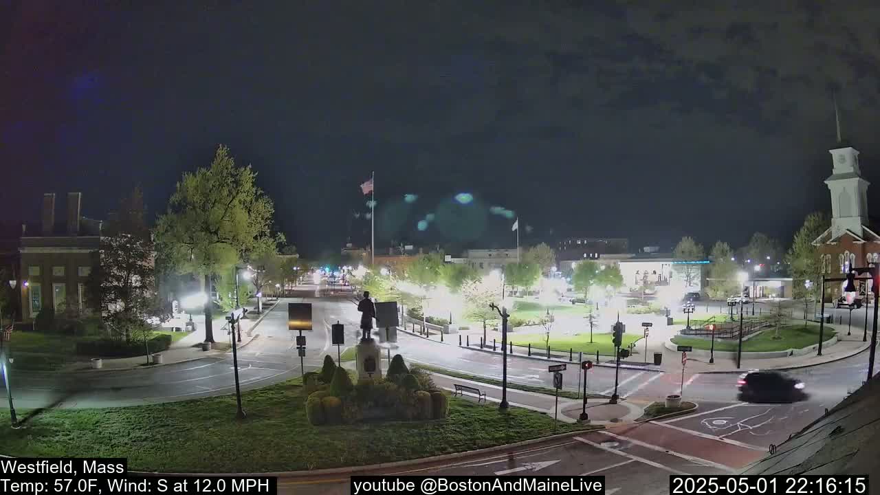 A nighttime view of a town square with a statue, surrounding buildings, and a few cars driving on the roads under a dark, clear sky.