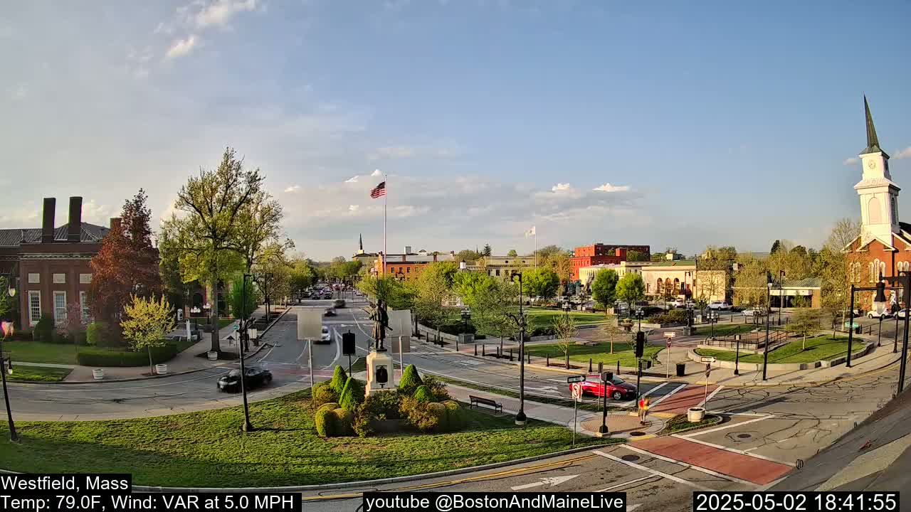 A partly sunny town square with brick buildings, green trees, and a monument, under a mostly clear sky.