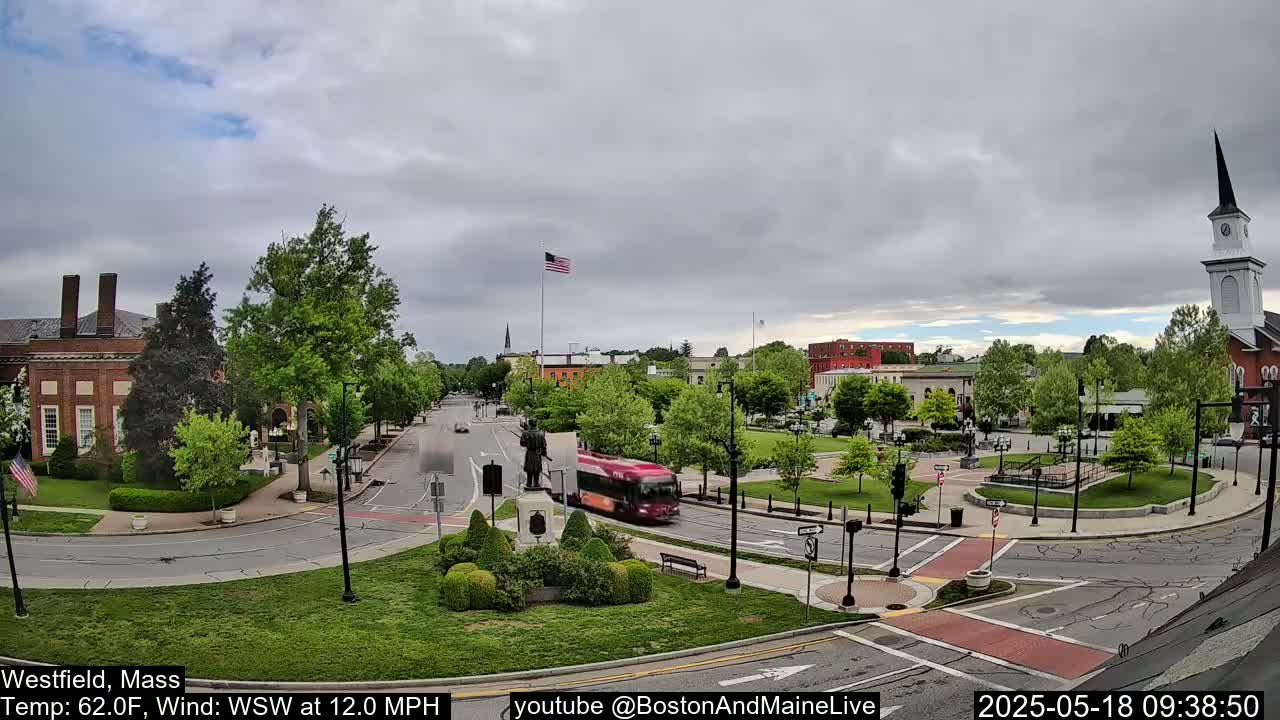 A town square with a red bus driving through it under an overcast sky.