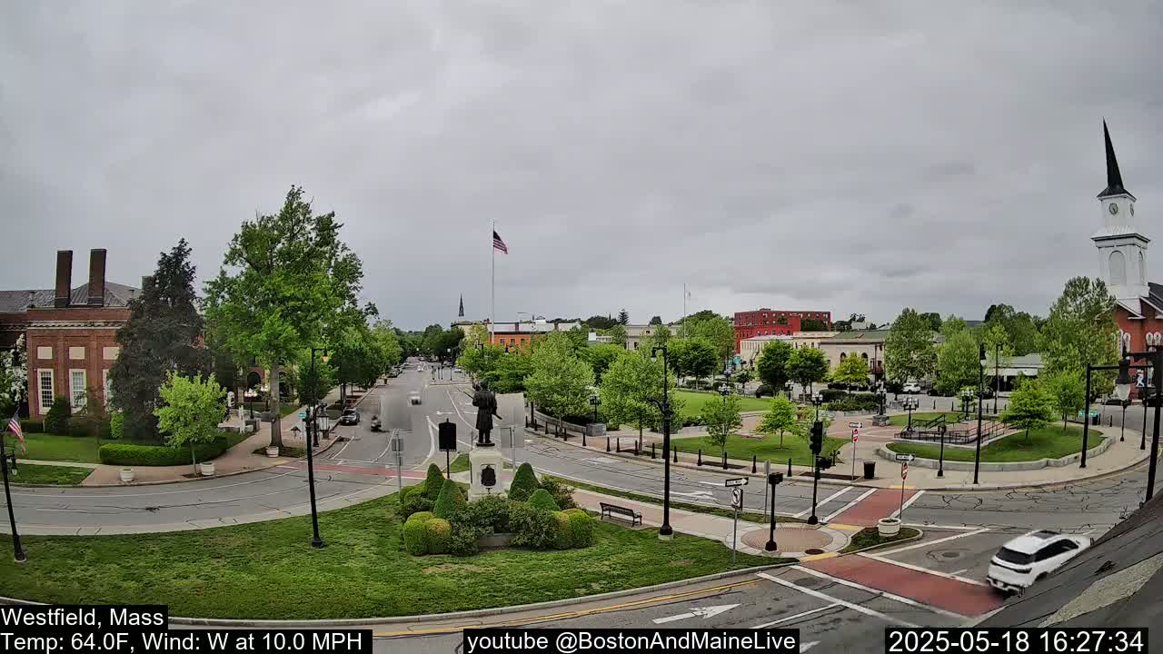 A town square with a statue and a church, under a cloudy sky, with cars driving on surrounding roads.