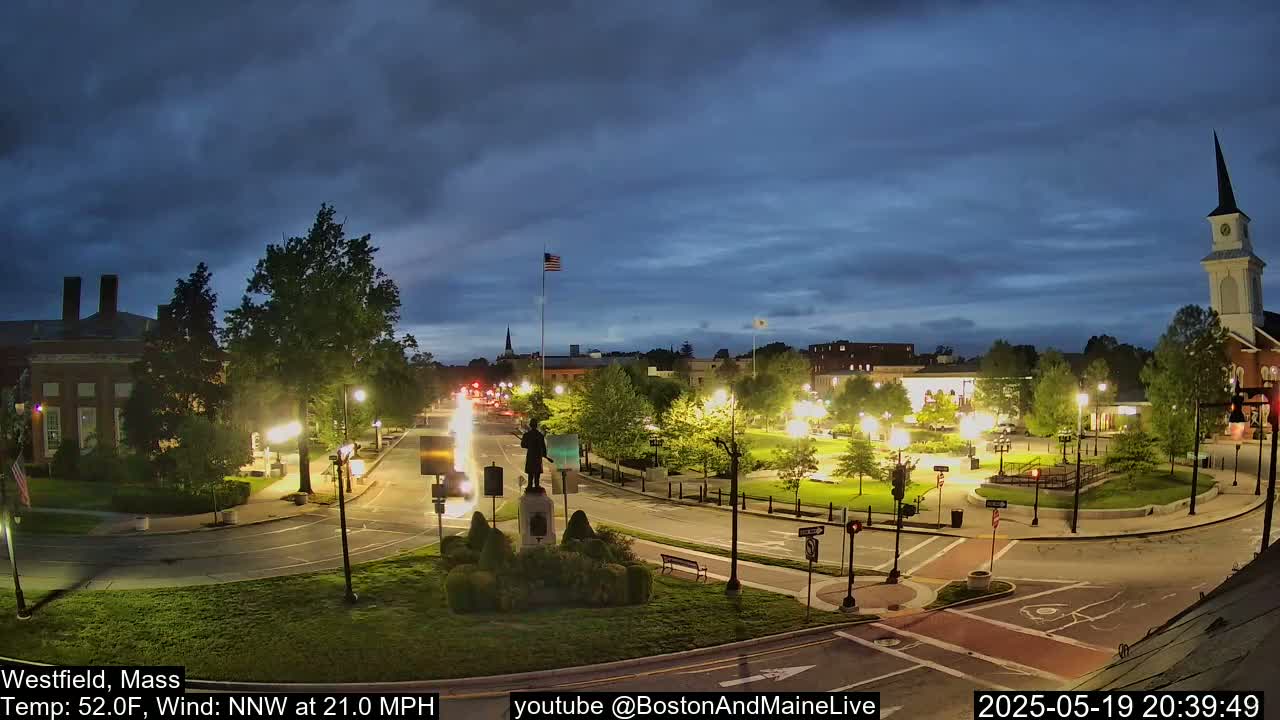 A town square at dusk, lit by streetlights, with a statue in the center, under a partly cloudy sky and a 21 mph wind.