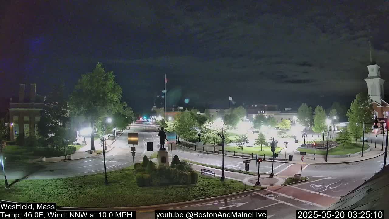 A nighttime view of a town square with a statue at its center, illuminated by streetlights under a partly cloudy sky.