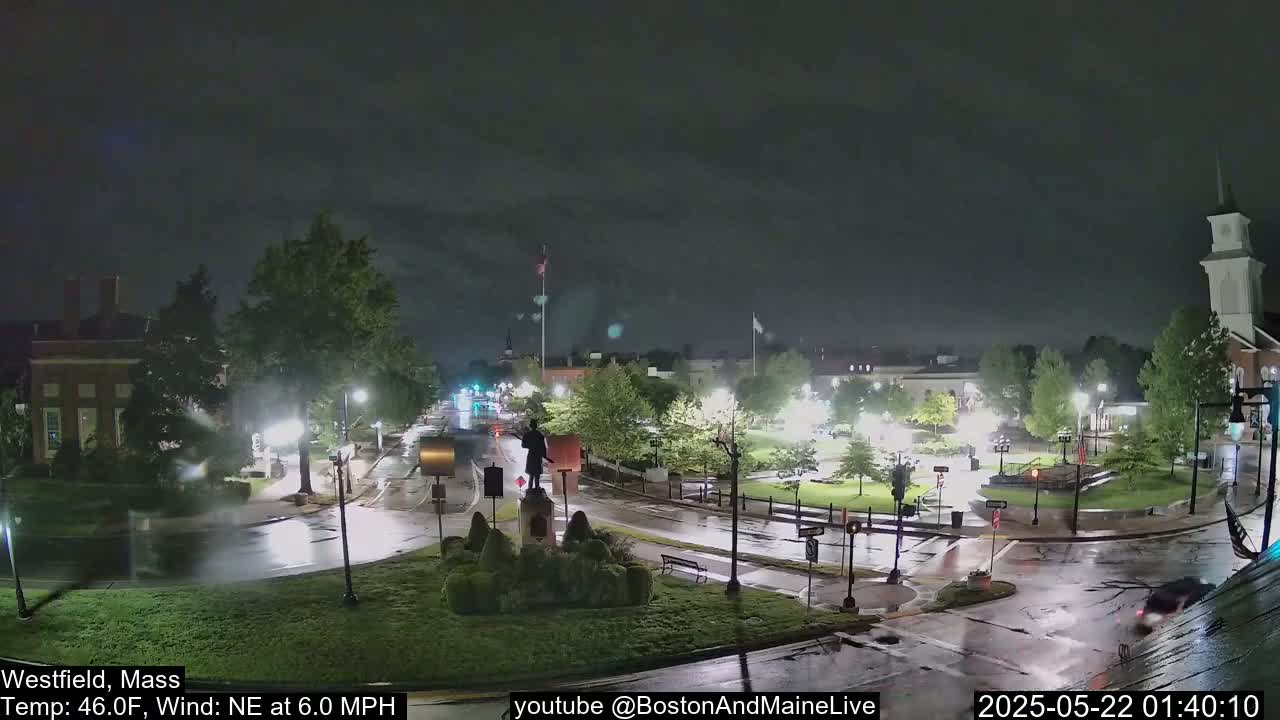 A nighttime view of a town square with a statue in the center, wet roads, and a few cars, under a dark, cloudy sky.
