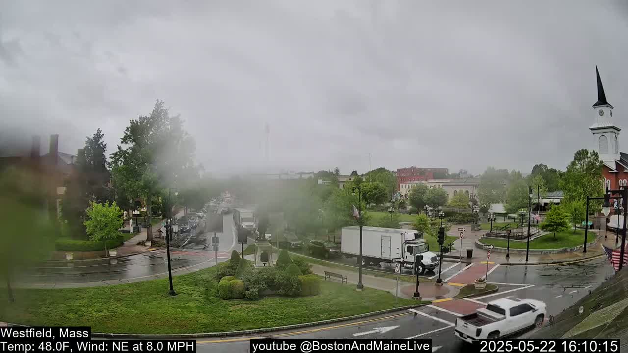 A town square with a church steeple in the background is seen on a cloudy, rainy day with traffic moving through the streets.