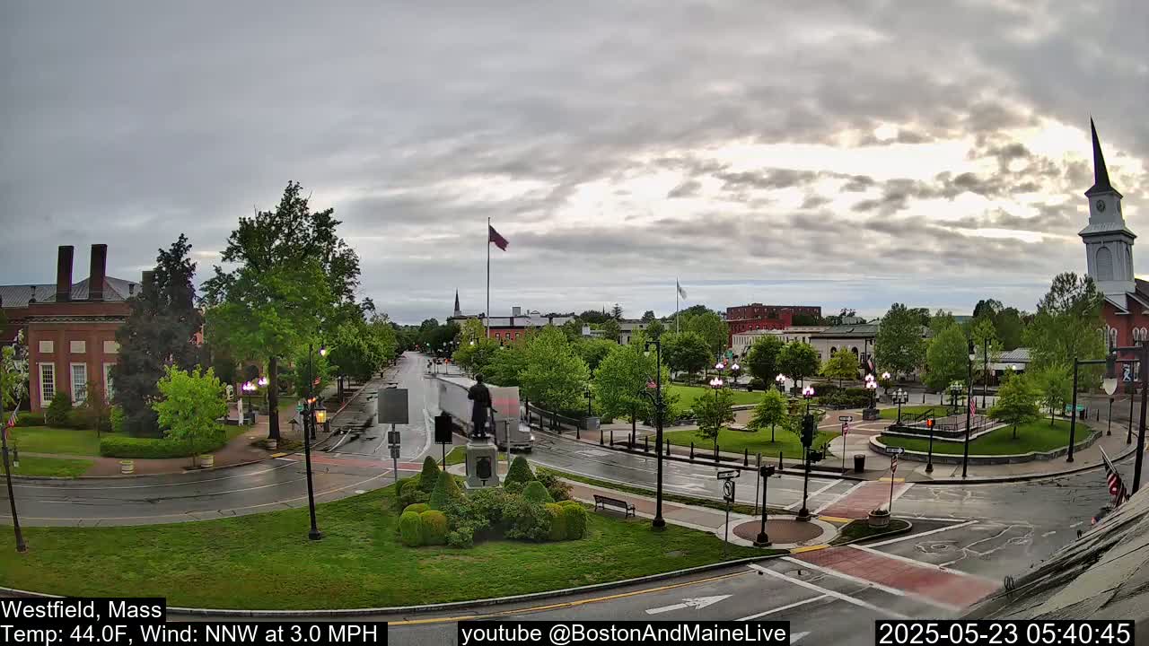 A town square with a monument and church under a cloudy sky, with light rain on wet streets and a temperature of 44°F.