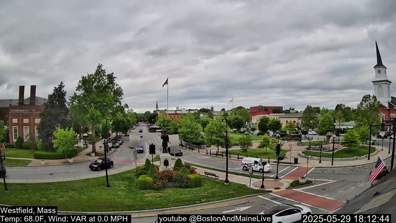A town square with a monument, several buildings, and green spaces under an overcast sky.