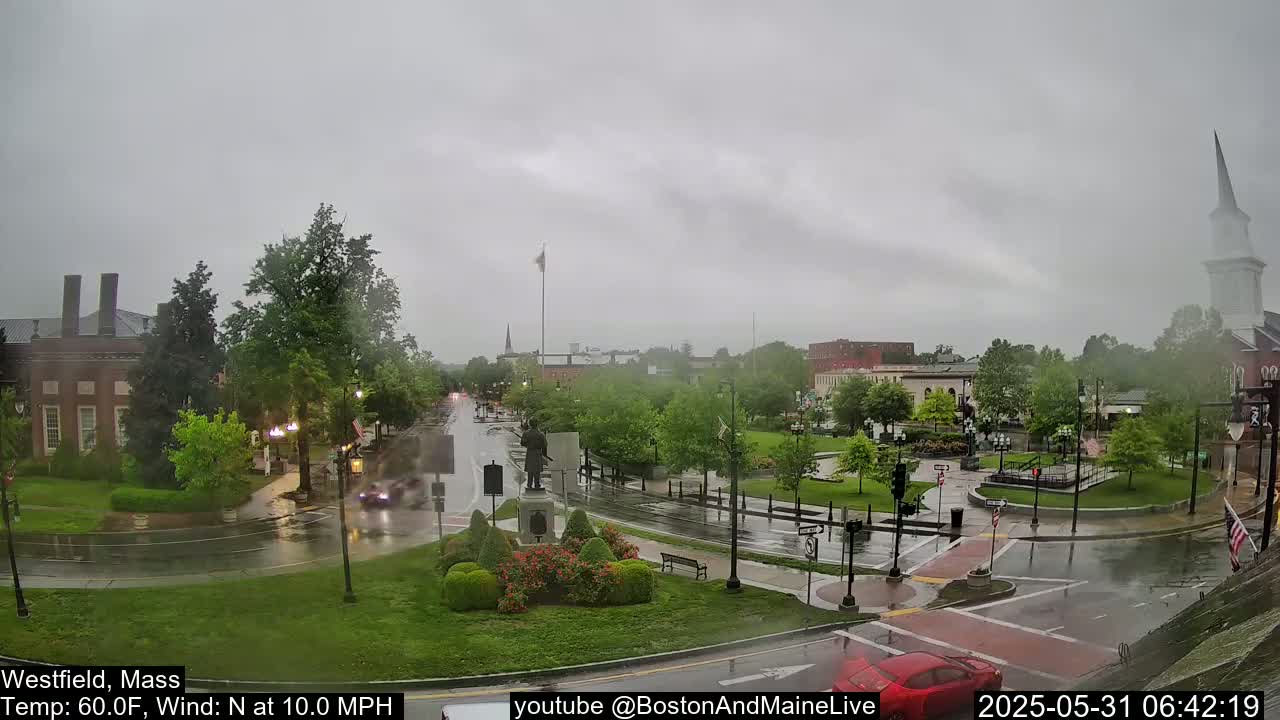A town square with wet streets and a monument in the center under an overcast sky, with a light rain falling.