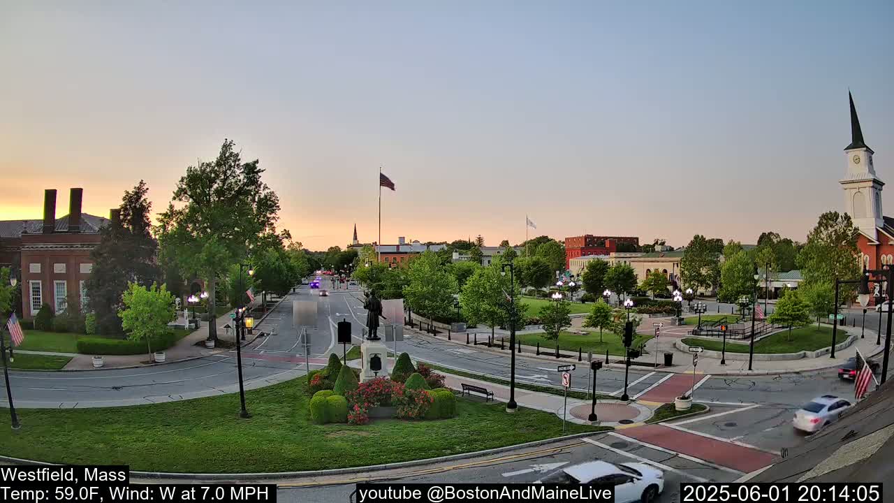 A quiet town square at sunset, with a few cars on the road and a statue in the center, under a clear sky.