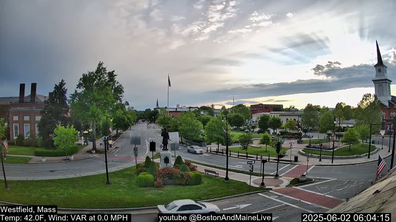 A mostly cloudy sunrise illuminates a town square with a statue, several buildings, and cars driving on roads.