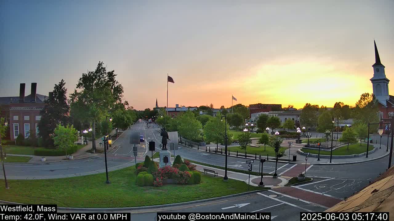 A town square at sunset, with a statue in the center, surrounded by buildings, trees, and a church steeple in the background, under clear skies.