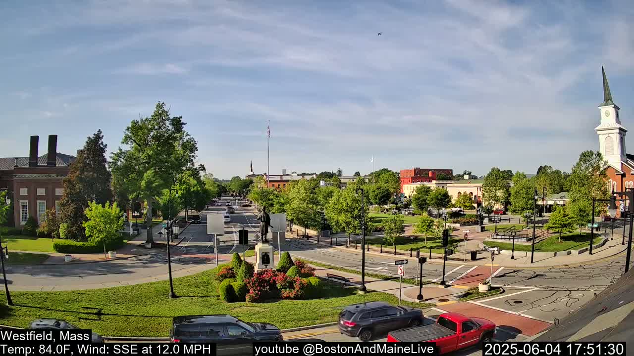 A town square with a statue and a church in the background under a partly cloudy sky.