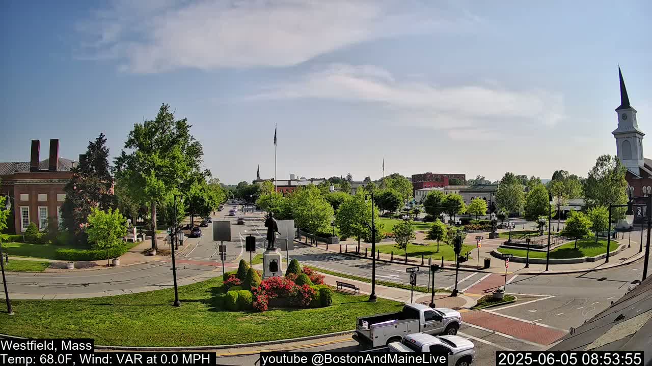 A partly sunny day reveals a town square with a statue at its center, surrounded by low-lying greenery and brick buildings, with a church steeple visible in the distance.